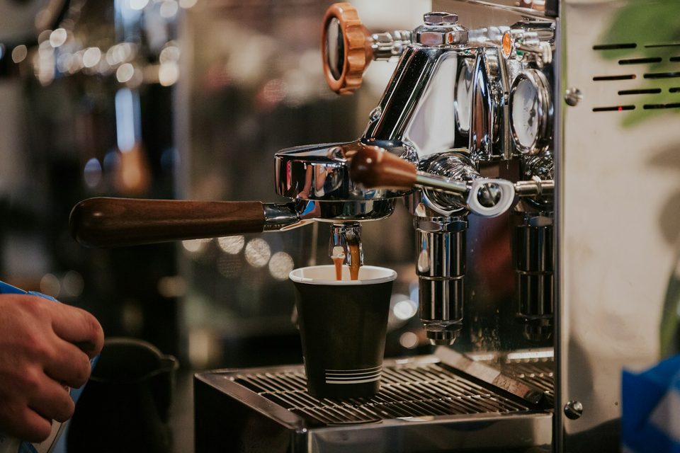 Chrome espresso machine pulling a shot into a cup, wood portafilter handle in the foreground.